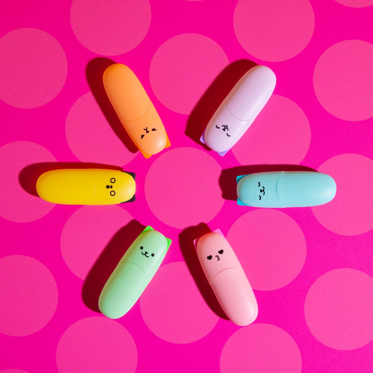 Six pastel kitty cat highlighters arranged in a circle on a pink polka dot background.