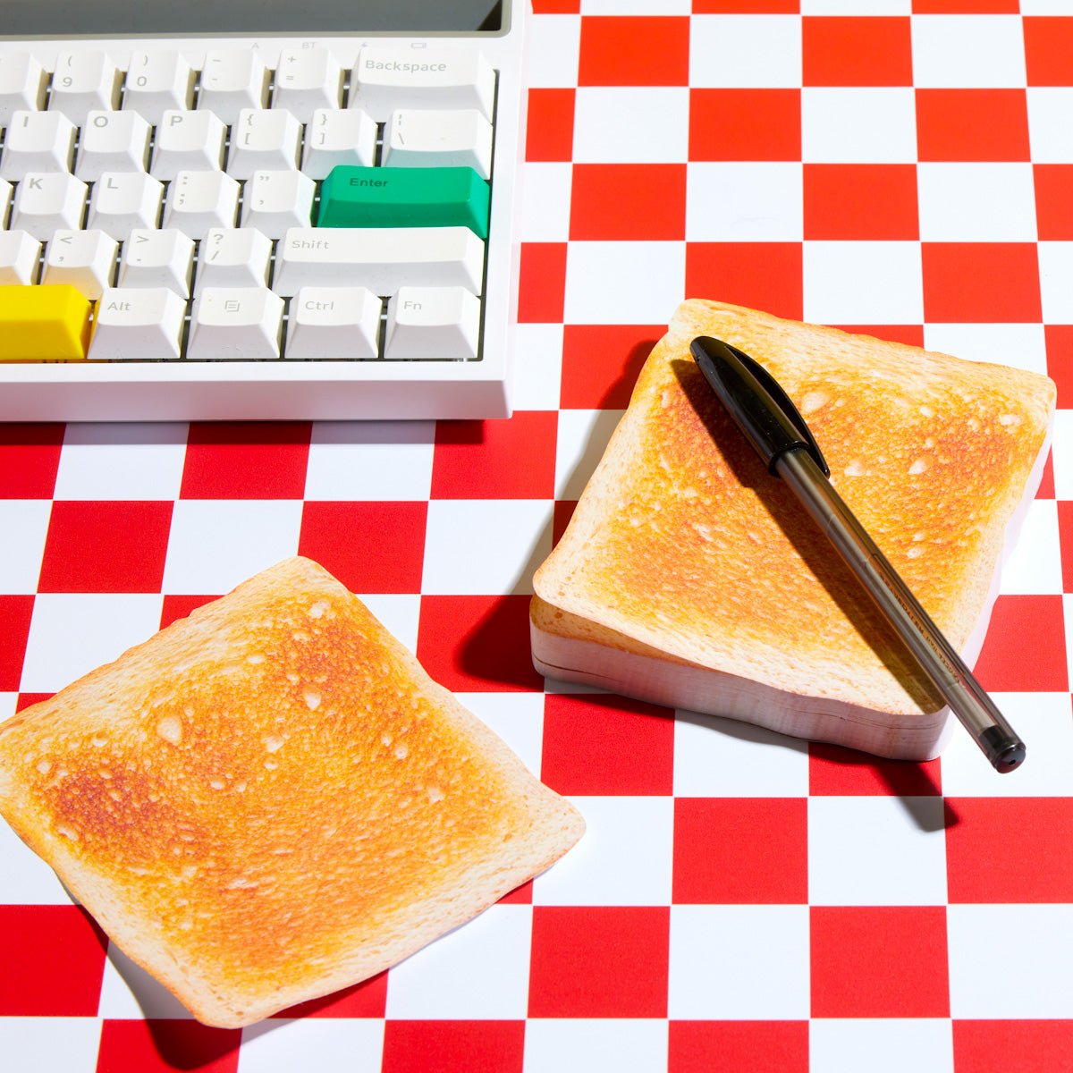 Toast-shaped sticky notes with a pen on a red checkered desk beside a computer keyboard.