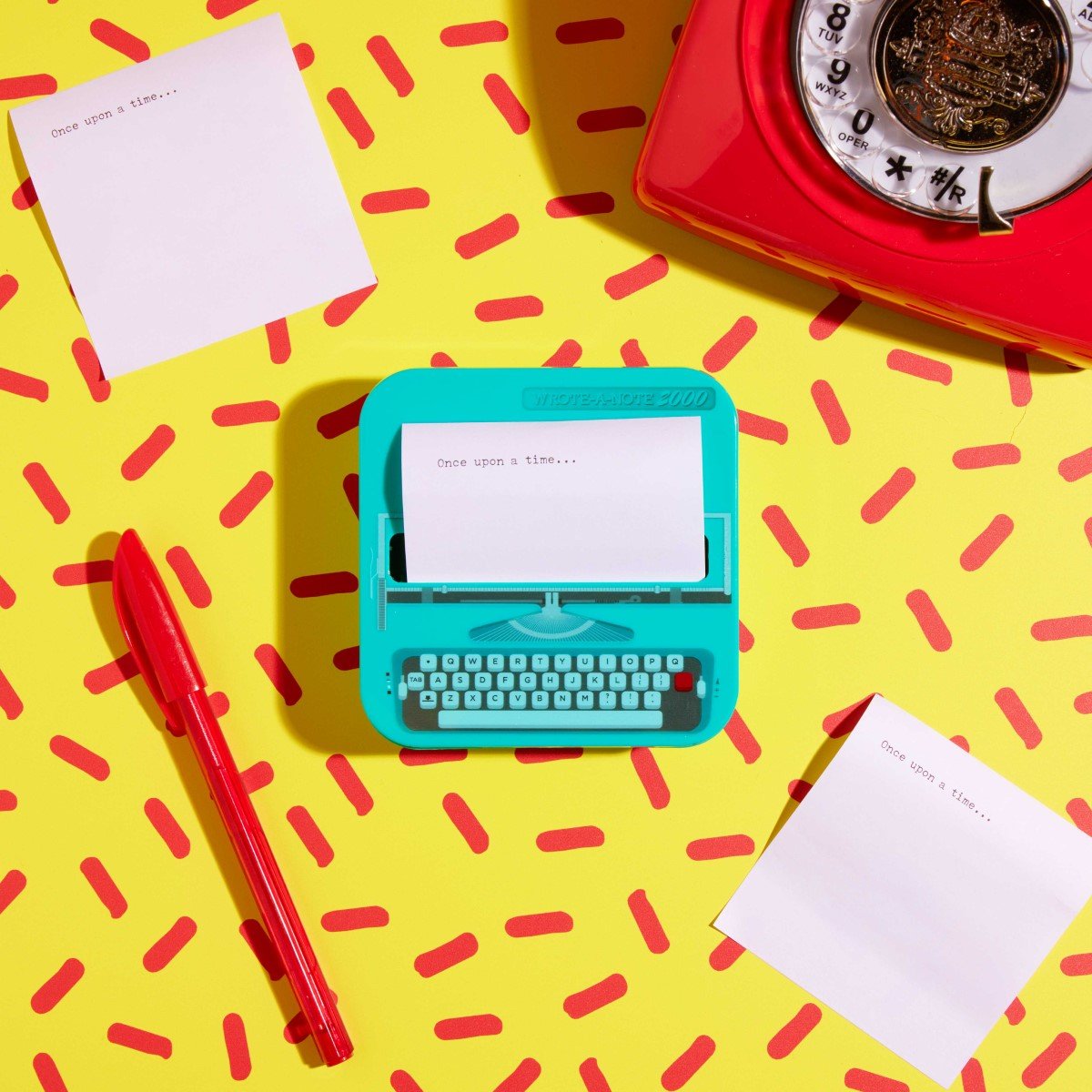 Retro typewriter-shaped sticky note dispenser on yellow patterned desk with notes and pen.